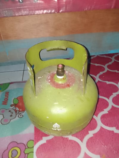 A green gas cylinder is displayed on a patterned pink and white mat. The cylinder is upright and positioned slightly off-center to the right. It has a yellow handle at the top. The mat has a repeating quatrefoil pattern in white on a pink background. To the left of the cylinder, a portion of a mat with a cartoon mouse and the word "Mouse" is visible. The background consists of a wooden surface above a light blue surface. The lighting suggests an indoor environment. The image is likely taken in Cisarua, Indonesia, given the common use of such gas cylinders in households there. There are no people or visible actions in the image.