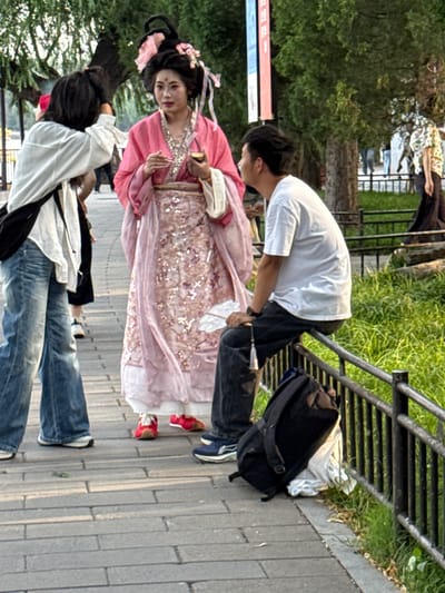 The image depicts a scene in Xicheng District, China, possibly within a park or pedestrian walkway. In the foreground, a person wearing a white shirt and blue jeans is walking away from the camera. To their right, a person in traditional Chinese attire, including a pink dress and elaborate headwear adorned with flowers, is standing and holding an object. Next to the person in traditional attire, a person in a white t-shirt and dark pants is sitting on a railing. A black backpack is placed on the ground near the person sitting on the railing. The background shows trees, grass, and a black metal fence. Several other individuals are visible in the background, some walking and others standing. The ground is paved with rectangular stones.