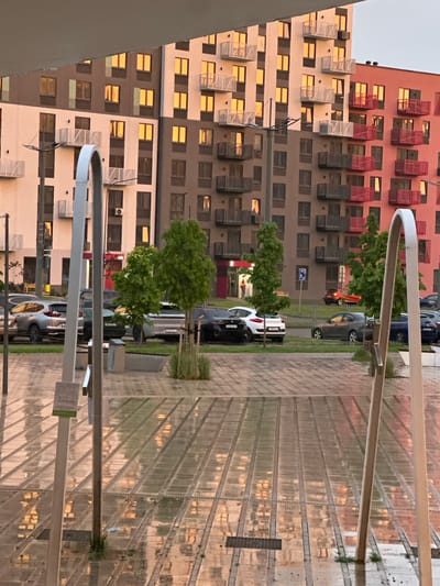 The image captures an urban residential scene in Бараўлянскі сельскі Савет, Belarus, characterized by low-angle warm sunlight or artificial illumination reflected on various surfaces. The foreground displays a wet, paved plaza featuring parallel translucent or reflective panels. Two prominent, light-colored, arching metallic structures rise vertically from this surface, framing the central view. A pole with a small green-edged sign stands behind the left arch.
In the midground, a street runs horizontally, populated by several parked cars including a silver SUV, a white compact car, a dark blue sedan, and a red hatchback. Multiple young trees with green foliage are planted along the street and in landscaping beds. A blue pedestrian crossing sign is visible on a pole. A single person is faintly discernible inside a building entrance with a red frame on the left side of the street.
The background is dominated by multi-story apartment buildings. The building on the left features a facade with alternating light beige and dark brown sections, equipped with numerous balconies presenting white metal railings. The building on the right exhibits a reddish-pink facade with balconies featuring dark red railings. Nearly all visible windows in both buildings reflect a consistent, intense orange-yellow light. The upper portion of the background buildings is partially obscured by an overhead structure or awning extending into the frame. The sky above is a uniform light hue.