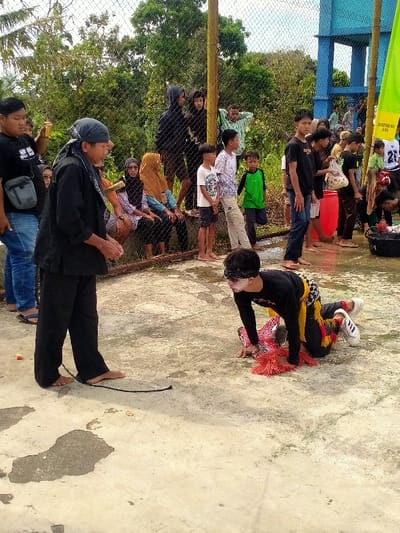 The image depicts an outdoor cultural performance or ritual occurring on a concrete surface in Tamansari, Indonesia. In the foreground, two male individuals are the central focus. An older man on the left, barefoot and dressed in black traditional attire including a head covering, stands with his gaze directed toward the right. A thin black rope or strap rests on the ground near his bare feet. To his right, a younger man is positioned on his hands and knees, wearing a black long-sleeved shirt, black and yellow patterned trousers, white sneakers, and a blindfold across his eyes. White makeup or paint is visible on his face. Red, fringed adornments are prominent around his waist and hands.
In the midground, a diverse group of spectators, including adults and children, observe the scene from behind a chain-link fence. Some individuals are seated on a low barrier behind the fence, while others stand. Many are dressed in casual attire; several women wear hijabs. The background features dense green foliage and trees, indicating a natural or semi-rural setting. To the far right, a tall blue water tank structure and a bright green flag attached to a pole are visible. The concrete ground in the foreground shows wet patches and scattered small orange objects. A black bucket is present on the right edge of the frame, near the spectators.