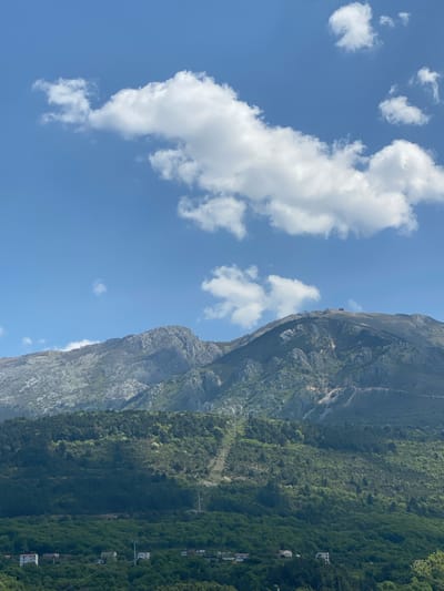 The image captures a vast landscape featuring a towering mountain, lush vegetation, and a clear blue sky with white clouds. The perspective is tilted, making the mountain appear on the right side, rising steeply from the bottom to the top of the frame. The sky occupies the left half and upper portions of the image, displaying a bright, clear blue hue. Scattered across the sky are several fluffy, white cumulus clouds of various sizes.

The mountain face is rugged and rocky, with visible geological contours. Its lower slopes are densely covered in vibrant green foliage, indicating a rich forest. Towards the base of the mountain and extending into the mid-ground, more greenery is visible, interspersed with what appear to be small residential buildings or structures, suggesting a settlement nestled within the natural environment. Thin lines, possibly power lines or cable car cables, traverse the forested area horizontally. A small, dark structure, perhaps a building or a beacon, is barely discernible on the upper ridge of the mountain against the bright sky.

The scene depicts a sunny day, likely late morning or early afternoon, given the bright, direct sunlight and distinct shadows on the mountain. The weather is fair, with a scattering of clouds but no indication of rain. The majestic natural elements combined with the subtle presence of human habitation suggest a location like Kavač, Montenegro, known for its mountainous terrain and picturesque coastal or valley settlements. No visible text is present in the image.