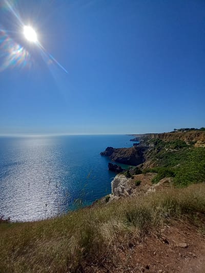 A wide-angle photograph captures a sun-drenched coastal landscape under a clear blue sky. The sun, partially obscured by lens flare, is positioned in the upper left corner, casting a bright glare across the scene. The foreground is dominated by dry, golden grass and reddish-brown soil, suggesting a dry climate. A path or trail is visible, winding through the vegetation.
The midground features a dramatic cliff face, with layers of rock in shades of brown and white. Lush green vegetation clings to the upper slopes of the cliffs. Rocky outcrops rise from the turquoise water that laps against the base of the cliffs.
The background shows a vast expanse of calm, deep blue ocean stretching to the horizon, where it meets a hazy, lighter blue sky. The ocean surface is textured with shimmering reflections of sunlight. Along the distant coastline, the cliffs continue, with hints of buildings or structures visible on the clifftops. The overall impression is one of a rugged, sun-drenched coastline, likely a natural preserve or park.
This specific scene does not depict any elements that are characteristic of the city of Lima, Peru. Lima is a coastal capital city known for its urban sprawl, colonial architecture, and a generally different type of coastline, often characterized by a more developed urban edge and beaches that are more frequented by people. The rugged, undeveloped cliffs and dry grasses in this image are not typical of Lima's known coastal areas.