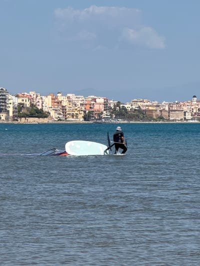 The image shows a person windsurfing in the sea near the city of Anzio, Italy. The person is wearing dark clothing and a light-colored hat. The windsurf board is white, and the sail has red and black accents. The buildings of Anzio line the coast in the background. The sky is blue with a few clouds.