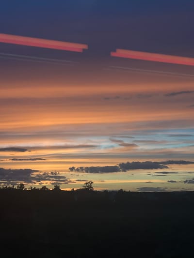 The image depicts a sunset with distinct horizontal cloud formations. The sky transitions from a deep blue at the top to shades of orange, peach, and pale blue towards the horizon. Two elongated, bright pink streaks are visible in the upper part of the sky, appearing as light trails or reflections. Below the colorful sky, a dark silhouette of a treeline and hills spans the lower portion of the image, obscuring any details of the ground or foreground. The scene is devoid of any visible people or specific landmarks that would pinpoint a location within Baiersdorf, Germany.