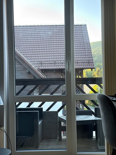 The image shows a view through a window of an outdoor area with a wooden deck and some furniture. On the left side of the image, there is a close-up view of a dark-tiled roof. The scene appears to be from a residential or vacation setting.
In the upper section of the window, we see a portion of a roof with dark, wavy tiles. Beyond the roof, a lush green forest or hillside is visible under a bright, possibly morning or afternoon sky. There are some wooden structures and railings on a deck. In the upper right quadrant, several pieces of dark, woven outdoor furniture are stacked, including what appear to be chairs or lounge seating. The furniture has a wicker-like texture.
The lower section of the window provides a different perspective on the same area. A larger section of a dark-tiled roof is visible, extending horizontally across the frame. A wooden-clad wall of a building is also present, with diagonal wooden beams forming a railing or structural element on a deck. More dark outdoor furniture is visible in the lower right quadrant, arranged on the deck. The furniture is dark and appears to be made of a woven material.
The scene suggests an outdoor living space, possibly a balcony or patio, attached to a building with a distinctive tiled roof. The lighting indicates it is daytime, and the bright sky suggests fair weather. The location context provided, Szklarska Poręba, Poland, places the scene in a mountainous or hilly region known for its natural beauty, which is consistent with the forested background. No people are visible in the image.