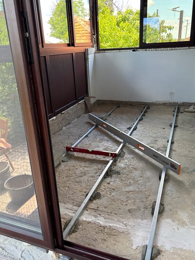 The image depicts an interior construction scene, possibly in a building in Pchelnik, Bulgaria. The focus is on a rough concrete wall being prepared for finishing. Horizontal metal channels are attached to the wall with what appears to be mortar or adhesive. Two levels, a "Gadget" 2000mm and a "Parkside" level, are positioned against the metal channels, likely being used to ensure they are aligned and level. A partially visible brown window frame is in the foreground. Outside the window, green trees and a blue sky can be seen. Inside, the foreground reflection also shows buckets and possibly a hand.