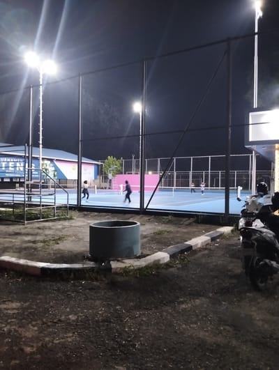 This media file captures an outdoor tennis court at night, illuminated by bright floodlights. The court itself is a vibrant blue, with white lines marking the boundaries. A net divides the court, and two individuals are actively playing tennis. One player, wearing dark clothing, is positioned on one side of the net, while another player, in lighter attire, is on the opposite side.
In the background, a building with a blue roof and colorful banners is visible. Text on one of the banners reads "TURNAMEN TENIS". The surrounding area appears to be a mix of asphalt and dirt, with some grass and foliage. A large cylindrical concrete structure is situated in the foreground, near the edge of the court. To the right, two motorcycles are parked, one prominently in the foreground.
The time of day is clearly night, indicated by the darkness of the sky and the artificial lighting. The weather appears clear. The scene evokes a sense of active recreation and sportsmanship, with the players focused on their game. The presence of the building with banners suggests that a tournament or event is underway. The location context is Palohgadeng, Indonesia.