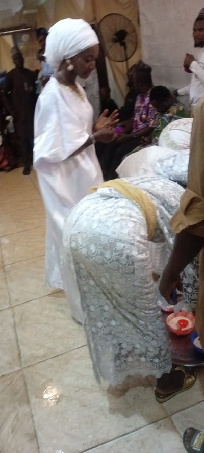 The image depicts an indoor scene in Dan Maliki, Nigeria, likely a social gathering or ceremony. The subjects are primarily people, with a focus on two women in the foreground. One woman, in the middle ground and to the left, is wearing a white hijab and a flowing white garment, possibly traditional attire. She is looking down and appears to be engaged in an activity, with her hands reaching out. Her expression is serene. Behind her, several other individuals are visible, some seated and others standing. Another woman in the foreground, to the right, is wearing a light-colored, lacy dress and is bent over, also appearing to be involved in an activity. Her face is not clearly visible.
In the immediate foreground, on a tray, are several small bowls. One red bowl contains a white substance with a red utensil, suggesting food or a cosmetic product. The floor is tiled with a light brown and beige pattern. In the background, a fan is visible on the wall, and the lighting suggests it might be daytime or an evening event with artificial lighting. The overall atmosphere seems calm and possibly celebratory. The clothing styles suggest a cultural event, possibly a wedding, naming ceremony, or religious gathering, given the prevalence of modest attire and head coverings. There is no visible text in the image.