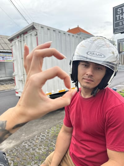 A medium shot captures a man wearing a white helmet and a red t-shirt. He is looking towards the camera, seemingly outdoors. A hand is positioned in the foreground, forming an "OK" gesture. The background includes a white truck, a building with a brown roof, and a street sign that is partially visible with the word "STOOLS." The location is Munggu, Indonesia. The image is oriented vertically.