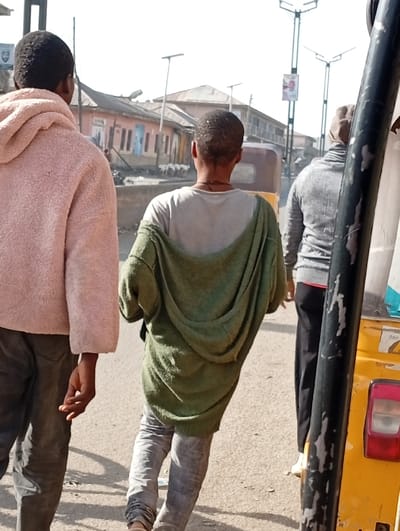The image depicts a daytime street scene in Gawuna, Nigeria. Three individuals are walking away from the viewer on a paved road. The leftmost person wears a pink, fuzzy hooded sweatshirt and dark jeans. The central person wears a light grey t-shirt, an olive green hooded sweatshirt draped over their back, and light grey patterned jeans. The person on the right, partially obstructed by a yellow auto-rickshaw, wears a grey long-sleeved top, dark pants, a light brown head covering, and light-colored footwear. A black, worn structural bar of the auto-rickshaw is visible on the far right. In the background, a line of single and multi-story buildings, some with light pink or muted facades and blue-painted windows, extends along the street. Multiple modern streetlights with integrated solar panels are present, one featuring a red and white banner. A group of white goats is visible in the middle distance on the left side of the street near the buildings.