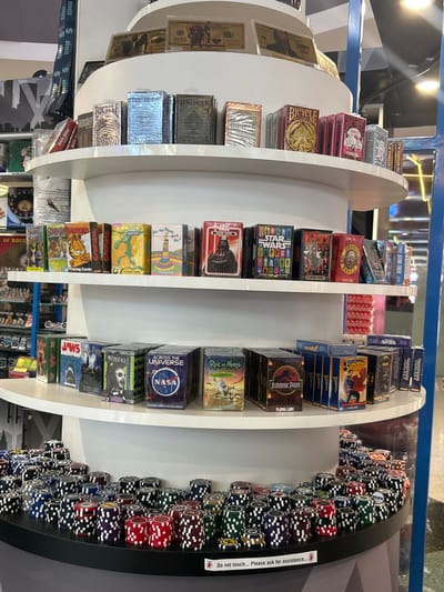 The image shows a multi-tiered display stand in what appears to be a retail environment, likely a shop or store in Las Vegas, United States. The stand holds various decks of playing cards featuring licensed properties such as Star Wars, NASA, Jurassic Park, and Marvel Comics characters. Poker chips are displayed along the right side of the stand. A sign advises customers not to touch and to request assistance.