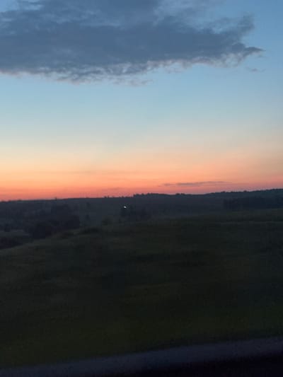 The image captures a serene sunset over a rural landscape in Estachi, Russia. The sky is painted with soft gradients of blue, light orange, and pink, transitioning into darker shades as it meets the horizon. A few scattered clouds drift across the upper portion of the sky. Below the sky, rolling hills covered in dark, lush vegetation stretch towards the horizon. A faint, small light is visible in the distance on the rolling hills, possibly a distant light source or a reflection. The foreground is dominated by dark, shadowy terrain, suggesting a field or meadow. The bottom edge of the image is bordered by a dark, possibly blurred, object, hinting at the viewpoint from which the photograph was taken. The overall scene conveys a sense of tranquility and natural beauty.