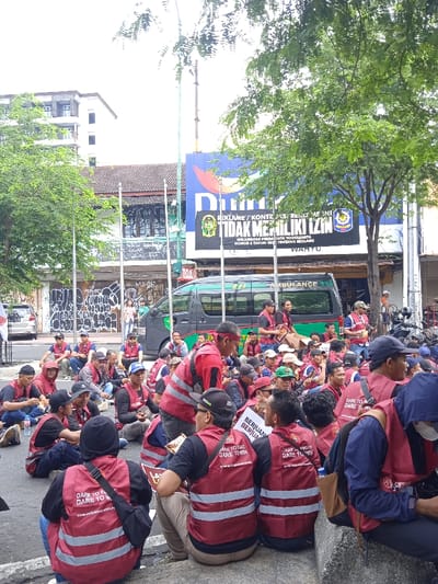 The image depicts a group of people in a large gathering on a street in Yogyakarta, Indonesia (latitude: -7.784, longitude: 110.367). The group is made up of mostly young men, wearing bright red vests with the slogan "DARE TO FIGHT DARE TO WIN" emblazoned on the back. Many are sitting or squatting on the street, while others are standing. They appear to be engaged in a demonstration or a protest. In the background, a large, black and white van emblazoned with the word "AMBULANCE" is parked on the side of the street. There is a large billboard to the left of the van, with a sign that reads "REKLAME / KONTRAK REKLAME INI TIDAK MEMILIKI IZIN" in Indonesian, which translates to "Advertising / Advertising contracts do not have permission." The scene is set on a busy urban street, with buildings and shops lining the street. There are several trees visible in the background, and the sky is partly cloudy.