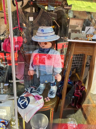 The image depicts a detailed retail store window display, located in Saint-Ouen-sur-Seine, France. A large, stylized mannequin, resembling a Playmobil figure with light skin, brown hair, and a smiling expression, is centrally positioned.
The mannequin is adorned with luxury fashion items. It wears a light blue wide-brimmed hat embellished with silver-colored beaded chains and blue Chanel 'CC' logos. A patterned blue scarf is tied around its neck. Its attire includes a layered top: a light blue base garment with a dark blue 'CC' logo pattern, covered by a pink, white, and light blue tweed-style jacket. A red camellia-shaped brooch is pinned to the jacket's left chest, alongside a small gold heart pendant on a chain. Light blue quilted denim-style shorts or a skirt cover the lower body. A small, light blue quilted Chanel crossbody bag with a gold chain strap and 'CC' logo is held in the mannequin's right hand. Its feet wear light grey ballet flats featuring a black and white camellia flower on the toe, over white ankle coverings or socks branded with "Chanel" and a 'CC' logo.
Behind the mannequin, a dark patterned Chanel cardigan, bearing a visible label and a red 'CC' brooch, hangs on a display. To the left, a pink quilted Dior saddle bag with a 'CD' charm is suspended. On the lower left, a white silk Chanel scarf with a prominent blue 'CC' logo and a pink border is folded on a gold, ornately carved stool. Chanel N°5 perfume bottles are also visible at the bottom.
To the right of the mannequin, a multi-tiered light brown wooden display stand holds a white Bvlgari wallet, assorted jewelry pieces (rings, brooches, a gold 'CD' logo charm), and a white shopping bag with a black ribbon, likely from Dior. Below the stand, large, reflective blue and red sculptures, resembling balloon animals, are visible. A partially obscured black and white poster depicting Marilyn Monroe is visible in the background. Reflections on the clear glass of the display window indicate an exterior urban environment.