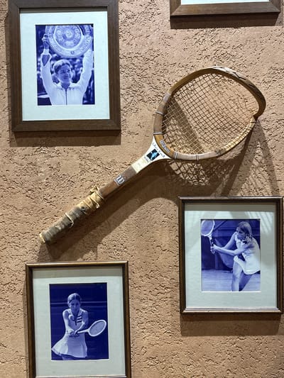 A wall display features a vintage wooden tennis racket and three framed, blue-toned monochrome photographs of female tennis players, all set against a textured, reddish-brown stucco wall. The specific location within Cukarica Urban Municipality, Serbia, cannot be determined from the image content.
The central object is an aged, light-colored wooden tennis racket, labeled "Wilson" and "Chris Evert" on its throat. The racket shows significant wear, including a damaged and cracked section on the upper right side of its head and a worn, fabric-wrapped grip secured by a visible screw and cord. It is mounted diagonally on the wall, extending from the lower-left to the upper-right quadrant.
Above the racket, a framed photograph depicts a smiling woman in a white tennis outfit, holding a large, ornate silver trophy (resembling the Wimbledon Venus Rosewater Dish) overhead with both hands. The background is blurred, suggesting an outdoor setting.
To the lower left of the racket, another framed photograph shows a woman in a white tennis dress with a focused expression, poised to execute a two-handed backhand shot.
To the lower right of the racket, a third framed photograph features a woman in a white tennis outfit in a low, athletic stance, holding her racket with both hands, appearing ready to strike the ball.
All three photographs are matted with a light blue border and enclosed in dark brown wooden frames. Distinct shadows are cast on the textured wall from the framed items and the tennis racket, indicating a light source from the upper-left.