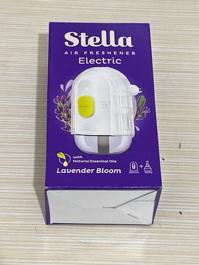 The image displays a purple cardboard box for an "Stella Air Freshener Electric" product, positioned horizontally on a light-colored, striped surface, likely a table or countertop. The box is seen from a slightly elevated angle, with its top and right side visible.

The front of the box is predominantly purple with white text and graphics. The brand name "Stella" is prominently displayed in a large, cursive white font on the left side. Below it, in smaller white text, are the words "AIR FRESHENER Electric." A graphic depiction of the actual white electric air freshener device, featuring a yellow adjustment switch, is central on the box, surrounded by illustrated lavender flowers.

On the right side of the box, text reads "with Natural Essential Oils" and below that, in a larger stylized script, "Lavender Bloom," indicating the fragrance. An icon depicting a leaf and a droplet is associated with the natural essential oils claim. In the top right corner, small white icons illustrate the product components: a plug-in device labeled "Device" and a refill bottle labeled "Refill," connected by a plus sign, signifying the combination.

The box appears to be in new condition, suggesting it contains an unused product. No people are visible, and no specific activity is taking place other than the static presentation of the packaged item. The lighting is uniform, indicating an indoor setting, likely during the daytime or under artificial illumination. The setting is a simple, uncluttered environment, consistent with a display or product shot. The image provides a clear view of the product's branding, type, and scent.