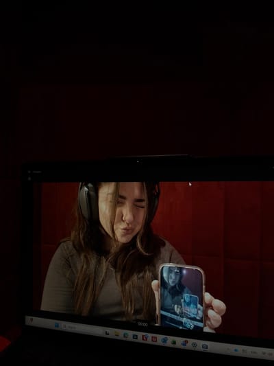 The image displays a close-up of a laptop screen, which is actively showing a live webcam feed of a woman. This scene is located in Bnei Brak, Israel.
**Laptop Interface:** The top bezel of the black laptop screen displays the text "Camera" and a small light indicator. The bottom portion of the screen shows part of the Windows taskbar, including a search bar, several application icons (e.g., Microsoft Edge, settings, photos), and a digital timestamp "00:00". A white mouse cursor is visible near the top right of the webcam feed area.
**Webcam Feed Content:** The main subject of the webcam feed is a young adult female with long, dark brown hair. She is wearing dark, over-ear headphones and a grey, long-sleeved shirt. Her facial expression is a puckered or "pouty" pose, with her eyes partially squinted. She is holding a smartphone in her left hand (viewer's right), extended towards the camera. The smartphone has a reflective, light-colored case. Its screen clearly displays a reflection of the woman holding it, with a smaller, recursive reflection of the phone itself visible within that reflection, demonstrating a Droste effect.
**Background (within webcam feed):** Behind the woman is a wall covered in dark red, rectangular panels, arranged in a grid pattern.
**Environment:** The ambient lighting around the laptop is dim, contrasting with the illuminated scene displayed on the screen.