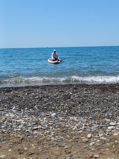 A person sits on a paddleboard in the ocean, holding a paddle. The person is wearing a white hooded jacket and dark shorts. The paddleboard is white with red accents. The ocean is blue with small waves, and the sky is clear and blue, suggesting a sunny day. In the distance, a small boat or ship is visible on the horizon.
The foreground of the image shows a beach covered in pebbles and sand. The pebbles are of various sizes and colors, from light grey to dark grey and brown. The sand is a light brown color. The waves are breaking on the shore, creating a foamy white line.
The scene appears to be a leisurely activity on a warm, clear day, likely during daylight hours given the bright sunshine and clear blue sky. The location is Krinitsa, Russia. There is no visible text in the image.