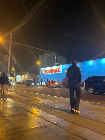 A nighttime street scene in Santiago de Surco, Peru, shows a person standing on a wet sidewalk facing a street with several cars. The person is wearing dark pants and a dark jacket, carrying a red bag with a pattern. Behind them, a large blue building with a bright red "SODIMAC" sign is visible. Several vehicles, including a red car and a dark SUV, are on the street. Streetlights and building lights illuminate the scene. The sidewalk appears to be made of concrete slabs. Power lines are visible overhead.