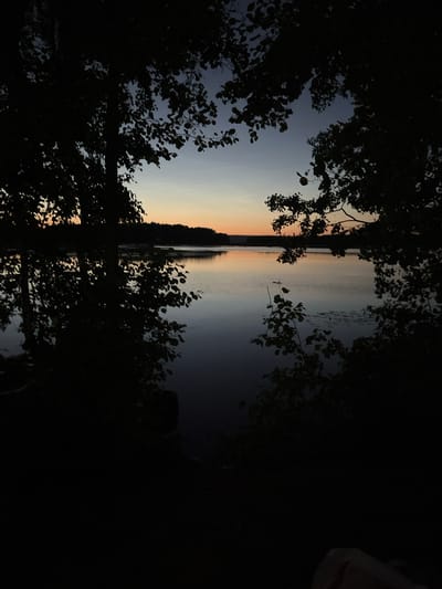This is a vertical, low-angle, outdoor shot of a lake at dusk, framed by dark trees. The sky exhibits a gradient of colors from dark blue at the top to orange and yellow near the horizon. The water is calm and reflects the colors of the sky. The foreground is dominated by the silhouettes of trees and branches, with some leaves visible. The overall mood is peaceful and serene. The location is identified as the city of Cheboksary, Russia. There are no people or specific objects of note in the scene other than the natural elements.