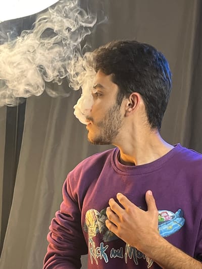 A man with dark hair and a beard exhales a thick cloud of white vapor from his mouth. He wears a purple sweatshirt with a colorful design on the front and rests his hand on his chest. The background is a plain gray. The light source seems to be coming from the left, illuminating the smoke and the man's face.