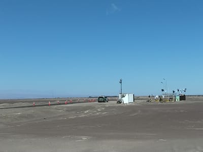 In Pozo Almonte, Chile, on a clear day, a desolate, dusty landscape stretches under a vast blue sky. A paved road runs horizontally across the middle ground, marked by a series of red and white traffic cones. A white vehicle, possibly a police car with green markings, is stopped on the road to the left of a small, white, modular building. Adjacent to this building is a smaller, brown structure with a simple roof, adorned with several black and white flags. A portable generator unit is parked near the white building. The scene is typical of arid, open regions, suggesting a remote location, possibly a checkpoint or a service area along a transportation route. The horizon shows distant, low-lying hills or mountains, and sparse vegetation dots the arid terrain.