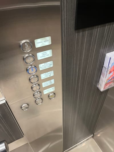 This image captures the interior of a modern elevator, focusing on its control panel and a section of the wall. The main subject is the stainless steel control panel, equipped with an array of circular buttons and associated rectangular labels. Above this panel, the elevator wall features a dark, horizontally striped wood-grain laminate. A small stack of brochures or pamphlets, partially obscured, rests on a ledge in the upper right.

No direct activity is occurring, but the illuminated "3" button suggests a floor has been selected. The scene depicts a clean, well-maintained elevator interior, likely within a commercial or retail complex like a shopping center, given the details on the labels. The location context of Indooroopilly, Australia further supports this.

Notable details include the design of the buttons, which have a sleek metallic finish, and the clear, functional labels. Visible text on these labels identifies destinations and parking levels:
- "PARKING - BROWN" (next to button 5)
- "CINEMA", "LIBRARY", "PARKING - PINK" (next to button 4)
- "MYER", "DAVID JONES", "FOOD COURT & DINING", "PARKING - GREEN" (next to button 3, which is illuminated)
- "TARGET", "PARKING - RED" (partially visible next to button 2)
- "COLES KMART", "GOURMET FRESH", "PARKING BLUE" (partially visible next to button 1)
- "PARKING YELLOW" (next to button P1)
Additionally, some text "ATRY" and "Directroy" is visible on the brochures. The overall aesthetic is contemporary and functional.