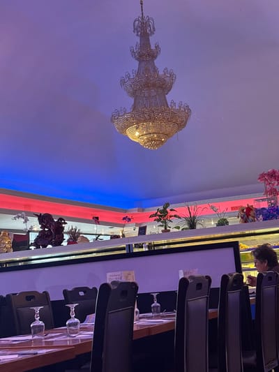 This image depicts the interior of a restaurant or buffet establishment located in Audun-le-Tiche, France. A large, ornate, multi-tiered golden chandelier with numerous crystals hangs centrally from a white ceiling. The upper perimeter of the room features recessed lighting with red LED strips on a higher level and blue LED strips on a lower, recessed ceiling section.
Below the ceiling, a long, white partition wall separates the foreground dining area from a service or buffet section. This partition includes a display shelf adorned with various decorative objects: a golden Buddha statue, a dark carved wooden figure resembling a mythical beast, multiple potted green plants, and pink and white artificial orchid arrangements. Behind this partition, on the right side, an adult female with short, dark, curly hair is seated at a table, wearing a dark top, and appears to be eating with a utensil in her right hand. Behind her, a glass display case consistent with a buffet or dessert counter is visible.
In the foreground, a row of unoccupied dining tables is visible, set with light-colored placemats and inverted wine or water glasses. Dark brown or black high-backed chairs are positioned at these tables. The lighting within the establishment combines the bright illumination from the chandelier with the vibrant colored accent lighting from the LED strips.