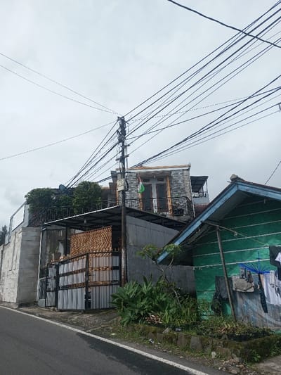 A street scene features an overcast sky over buildings. An abundance of power lines stretch across the upper portion of the image. The left side shows a gray wall and a metal gate. Further back is a stone-clad building with a balcony and a green flag. A small, green-painted shack stands on the right, adorned with hanging laundry and an umbrella. Vegetation is present in front of the buildings and along the street.
