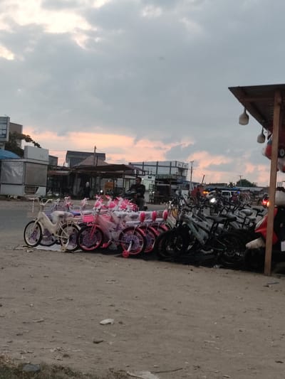 The image depicts an outdoor scene at dusk, likely in Meunasahaluedrienlhoksukon Barat, Indonesia. A row of bicycles, mostly pink and white, are lined up for sale or display on a dirt ground in the foreground. Behind the bicycles, various structures resembling shops or market stalls are visible, with some lights illuminated. A few individuals are present, including a person riding a motorcycle in the middle ground and another person standing near the shops. The sky is cloudy with a hint of orange and pink from the setting sun. The overall environment suggests a commercial area or a street-side market.