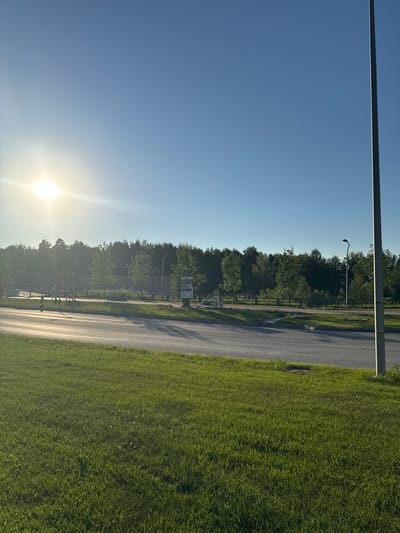 The image shows a sunny day in Koltsovo, Russia, with a bright sun low in the sky, casting long shadows. The scene is a roadside with a paved path running alongside a well-maintained grassy area. A tall lamp post stands vertically on the left side of the frame. Beyond the path, a dense line of green trees forms a backdrop. There are a few scattered trees within the treeline, and some structures, possibly signs or small buildings, are visible amongst the foliage. A person is walking on the path, and another figure appears to be sitting on a bench. The overall atmosphere is peaceful and serene, suggesting a quiet day outdoors. The clear blue sky and the vibrant green of the grass indicate pleasant weather.
