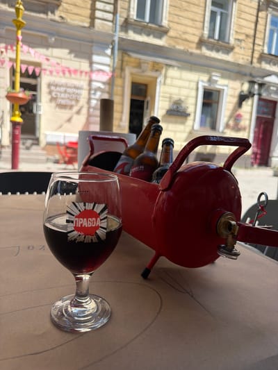 A glass of dark red liquid, likely wine or beer, sits on a table in front of a red, kettle-shaped object containing several beer bottles. The glass has a circular sticker with red and white radiating lines and text in Cyrillic script. In the background, a building with yellow walls and windows is visible. The scene appears to be an outdoor cafe in Ivano-Frankivsk, Ukraine. The building in the background likely houses a shop or restaurant, with signs in Cyrillic script indicating its purpose. A yellow lamppost with pink flags is also visible on the left side of the image, further suggesting an urban setting. The table surface has faint circular markings drawn on it, perhaps for a game or decorative purpose. The overall lighting suggests it is daytime.