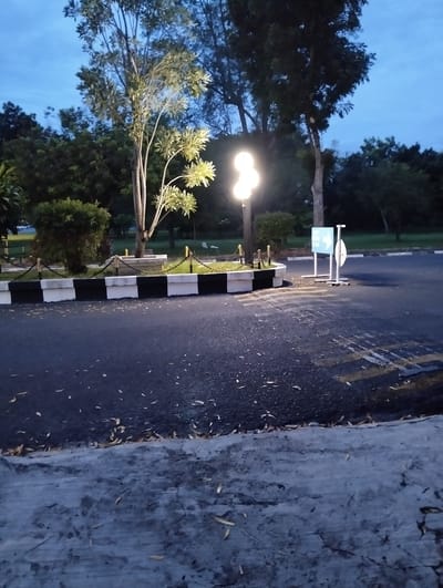 The image captures an outdoor scene, likely at dusk or dawn given the low light conditions. The setting appears to be a park or a landscaped area adjacent to an asphalt road. Several trees are visible, some with lush foliage and others more sparse, with a notable tree in the center that is illuminated by a streetlamp with multiple bulbs.
In the foreground, the asphalt road shows some wear and tear, with faint yellow markings and scattered dry leaves. A concrete barrier with alternating black and white painted sections runs along the edge of the road, separating it from a garden bed. This garden bed is enclosed by a low fence made of posts and chains.
To the right of the garden bed, a metal structure with a blue sign is present. The sign has some white text and graphics, including an arrow pointing to the left. The overall mood is quiet and serene, with the artificial light from the lamp casting shadows and illuminating parts of the scene. The weather appears clear, with a hint of blue in the sky. No people are visible in the image. The location context is Tambontunong, Indonesia.