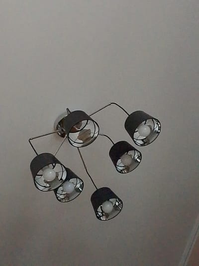 A multi-lamp ceiling light fixture hangs from a white ceiling. The fixture has a central chrome-plated base from which several metal arms extend, each holding a black lampshade. The lampshades are cylindrical with an open top and bottom, revealing a reflective interior and a white light bulb inside. The light fixture is positioned in the upper center of the image, against a plain white background that appears to be a ceiling. The image is taken from a low angle, looking upwards at the fixture. The location specified is Yanino-1, Russia, however, there are no visual cues in the image to confirm this location.