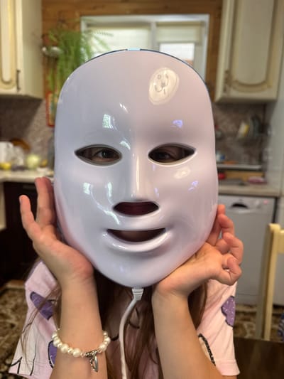 A young person is wearing a white LED facial mask. The mask covers most of their face, with openings for the eyes, nose, and mouth. The person's hands are placed on either side of the mask, supporting it. They are wearing a pink t-shirt with purple designs and a white beaded bracelet on their left wrist. The background appears to be a kitchen, with cabinets, a countertop, and a washing machine visible. There is also a plant in the background. The image is taken from a close-up perspective, focusing on the person's face and the mask.