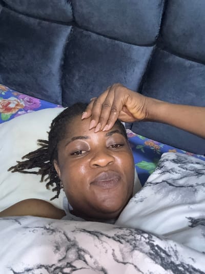 In a bedroom in Ajara, Nigeria, a close-up shot captures a woman lying on her side, propped up by a pillow. Her right hand rests on her forehead, and she is partially covered by a comforter with a marbled pattern. A floral patterned sheet is visible beneath the pillow. The background is dominated by a dark, tufted headboard.