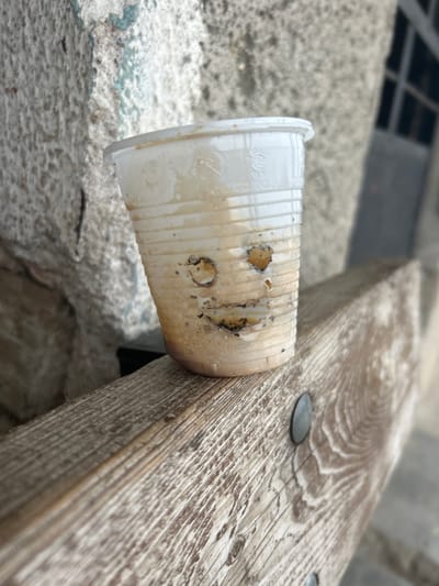 A translucent, ribbed plastic cup containing dried light brown liquid residue and sediment rests upright on a weathered, light-colored wooden surface in Dolni Tsibar, Bulgaria. The cup features three deliberate perforations on one side, configured as a face: two small, irregular circular holes for eyes and a larger, crescent-shaped hole forming a mouth. Faint embossed text and symbols are visible near the cup's rim. The wooden surface, characterized by pronounced grain and a bleached appearance, includes two dark, circular, flat fasteners. To the left, a rough, textured light gray wall is visible in the background. To the right, a blurred background reveals another light-toned building structure with a dark, rectangular architectural element.