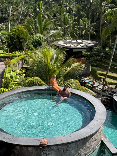 The image shows a couple in a circular pool, likely part of a resort or spa in a lush, tropical environment. The location is Tegal Suci, Indonesia. The woman is wearing an orange bikini. The man has a dark beard. Behind the pool are palm trees and other tropical vegetation. There is a large, decorative, black and white object in the background. Lounge chairs are placed near the staircase, further away from the pool.