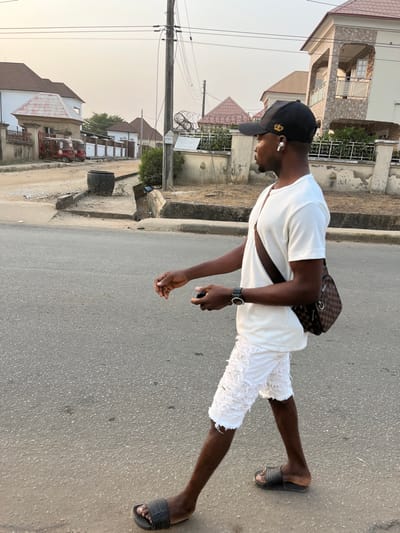 A man in a white t-shirt, tattered white shorts, and black slides stands on an asphalt road. He wears a black cap, a watch, and carries a checkered shoulder bag. He appears to be holding a phone or small object in his hand. Houses and power lines are visible in the background under a clear sky.
