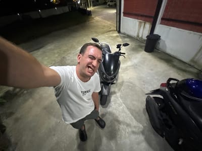 A man stands on a concrete driveway in front of a building. The man is wearing a white t-shirt with a graphic design, grey shorts, and black slip-on shoes. He is making a grimace and looking at the camera, which appears to be mounted on his scooter. To the man's right is a black and silver scooter. To the right of the scooter is another black scooter, partially visible. In the background, a black trash can is situated next to a white wall with red siding above it. The driveway is illuminated by artificial lighting, suggesting it is nighttime. The overall environment appears to be an urban or suburban setting, possibly a residential or commercial area. The location is identified as Wichit, Thailand.