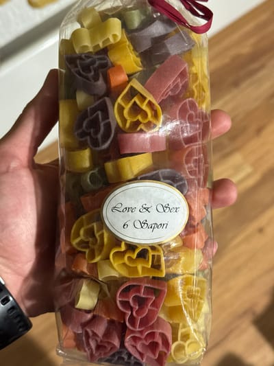 A person's hand is holding a clear plastic bag filled with heart-shaped pasta in various colors including yellow, red, purple, and orange. The bag is tied at the top with a dark red ribbon. A white oval label is adhered to the front of the bag. The label has a gold border and contains black text that reads "Love & Sex" and below that "6 Sapori". The background shows a wooden surface. The image is taken indoors, and there is no visible indication of the location in the city of Smolyan, Bulgaria.