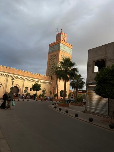 The image depicts a street scene in the Mechouar Kasbah area of Marrakesh, Morocco, under a cloudy sky. A tall minaret, adorned with patterned tile work, dominates the left side of the frame. Palm trees are visible in front of the minaret. Adjacent to the minaret is a long, crenellated wall enclosing a building. Along the street, people are present, some seated on benches and others walking. The street is paved and bordered by a line of spherical bollards. On the right, a building with a metal roller shutter is present. The overall scene conveys an urban landscape with architectural and cultural elements.