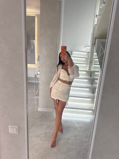 A full-body mirror selfie is taken by a young woman wearing a white two-piece outfit consisting of a ruched crop top with a lace-up front and long, sheer sleeves, and a matching ruched mini skirt with drawstring detailing. She is holding an orange smartphone in her right hand to take the photo. The woman has dark hair and is standing on a tiled floor.

The environment reflected in the mirror shows a modern interior with a prominent white staircase that is illuminated with embedded lights. The walls appear to be a light, textured material. To the left of the stairs, a portion of a mirrored hallway is visible, reflecting a console table with a lamp and some decorative items. A light switch is visible on the wall to the left of the mirror frame. The scene is well-lit, suggesting an indoor residential setting.