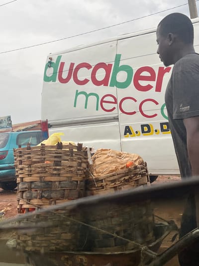 A person is leaning over a white truck with red and green lettering. The lettering appears to say "ducaberry mecca" and "A.D.D.". The person is wearing a grey t-shirt with some text on the sleeve. Behind the truck, there are two large woven baskets, one on top of the other, filled with what looks like produce. The baskets are made of bamboo or similar material and are tied with twine. A yellow plastic bag is visible between the baskets. A blue truck is partially visible behind the baskets, with a red object on top. The scene appears to be outdoors, with an overcast sky. The overall impression is that of a street market or a delivery scene in Sabojibi, Nigeria.