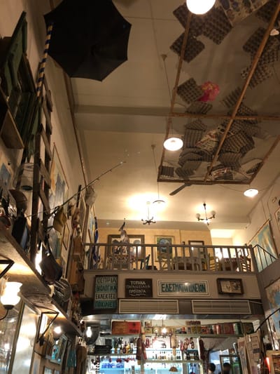 This image captures an indoor scene, likely a shop, market stall, or museum exhibit, filled with an eclectic assortment of objects. The perspective is looking down a long display counter, with a wall to the left and a deeper section of the space to the right. The lighting is artificial, provided by several bare lightbulbs hanging from the ceiling and wall-mounted fixtures, suggesting it could be evening or an establishment that relies solely on interior illumination.
In the foreground to the left, a closed black umbrella leans against a wall, next to a blue and white striped rope. A wooden box, possibly a tackle box, with green lining is open nearby. A fishing rod extends diagonally across the lower half of the frame, hinting at a potential theme of fishing or outdoors.
The main focus is a long, rustic-looking display table or counter, heavily laden with various items, including framed pictures, small sculptures, and miscellaneous curios. A wooden ladder-like structure is also part of this display. Behind the counter, a light-colored wall features framed artworks and a section of textured wallpaper.
Stacked vertically on this wall are prominent signs with Greek text. From top to bottom, they read:
1. "ΕΔΕΣΜΑΤΟΠΩΛΕΙΟΝ" (EDESMATOPOLEION), meaning "Delicatessen" or "Grocery Store."
2. "ΤΟΠΙΚΑ ΠΑΡΑΔΟΣΙΑΚΑ ΠΡΟΙΟΝΤΑ" (TOPIKA PARADOSIAKA PROIONTA), meaning "Local Traditional Products."
3. "ΟΠΩΡΙΑ ΒΙΟΛΟΓΙΚΑ ΕΚΛΕΚΤΕΣ ΠΟΙΚΙΛΙΕΣ" (OPORIA VIOLOGIKA EKLEKTES POIKILIES), meaning "Organic Fruits, Selected Varieties."
Further in the background, more shelves are visible, densely packed with merchandise. Above, a large, rectangular wooden frame covered with a net is suspended from the ceiling. Within this netting, numerous textured panels resembling egg cartons are arranged, alongside other decorative items, including a pink crumpled object. A ceiling fan is also visible.
No people are present in the image. The scene conveys a sense of a curated collection or a retail space specializing in traditional and local products, consistent with the given location of Marousi, Greece.