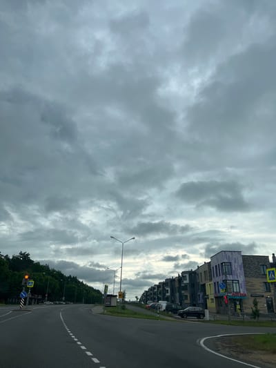 The image depicts a cloudy day in a suburban area. A paved road curves to the right, with white dashed lines marking the lanes. On the left side of the road, a traffic light shows a yellow signal, and a pedestrian crossing sign is visible. Further down, a densely wooded hill rises. Opposite the hill, on the right side of the road, is a row of modern multi-story buildings with varied architectural styles and colorful facades. Cars are parked along the buildings, and there are signs, including one for a pharmacy and another with a bicycle icon. Streetlights are positioned along the road. The sky is filled with thick, grey clouds, suggesting overcast weather.
