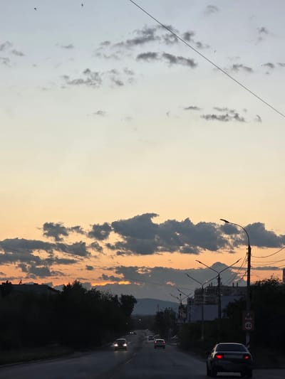 The image depicts a twilight scene on a road in Chita, Russia. The sky is a blend of pale blue and soft orange, indicating the late evening or early morning. Scattered dark clouds punctuate the sky, some edged with the warm glow of the setting or rising sun. A single, thin power line stretches diagonally across the left side of the frame, with a few distant birds visible in the sky.
On the right side of the image, a road stretches into the distance, bordered by dark trees and some buildings. A black car with its taillights illuminated is driving on the right side of the road, suggesting it is being driven on the right, which is unusual for Russia. A speed limit sign showing "40" is visible on the right side of the road. Several other vehicles are visible further down the road, their headlights on. The overall mood of the image is serene and somewhat atmospheric, capturing the transition between day and night.