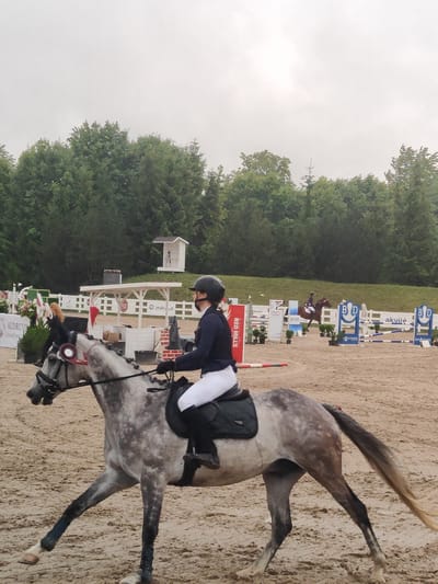 In Ziniūnai, Lithuania, a horse show is taking place. The image captures a dappled grey horse with a rider in a blue jacket and white pants, wearing a black helmet. The horse is in mid-stride on a sandy arena. In the background, there are various show jumping obstacles, including fences with blue and white poles. Other riders and horses are visible in the distance, as are trees and a cloudy sky. The scene suggests an outdoor equestrian event.
