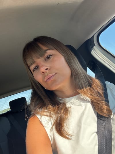 A woman is inside a car, likely seated in the passenger seat, as suggested by the angle and the presence of a headrest and seatbelt. The car appears to have a sunroof. The woman has brown hair with bangs, and she is wearing a white shirt. The car interior is a mix of gray and black. There is no direct evidence to definitively place this image in Saint-Maximin-la-Sainte-Baume, France. There are no visible landmarks, license plates, or other identifiers that could confirm the location.