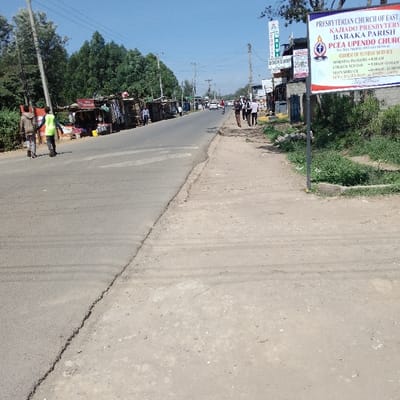 The image depicts a daytime street scene in Nkaimurunya ward, Kenya, under a clear blue sky. An asphalt road, exhibiting surface wear, extends from the foreground into the distance, appearing to curve slightly to the right. On the left side of the road, a continuous line of roadside vendor stalls, constructed from corrugated metal and wood, is present. Behind these stalls are dense green trees and utility poles with overhead power lines. Two individuals are observed walking on the asphalt road on the left, moving away from the viewer; one wears a bright yellow-green reflective vest. To the right of the asphalt road is a wide, unpaved dirt shoulder with patches of green vegetation. A large white billboard sign, bearing text for the "PRESBYTERIAN CHURCH OF EAST KAJIADO PRESBYTERY BARAKA PARISH PCEA UPENDO CHURCH," is prominently positioned on this right shoulder. Further along the right side, a group of approximately five to seven pedestrians is walking on the dirt shoulder, moving away from the foreground. Behind them and further down the road, more structured commercial buildings are visible, one with a sign indicating "BOOKSHOP." In the mid-ground and background, several vehicles are present on the road, and additional utility poles line the street.